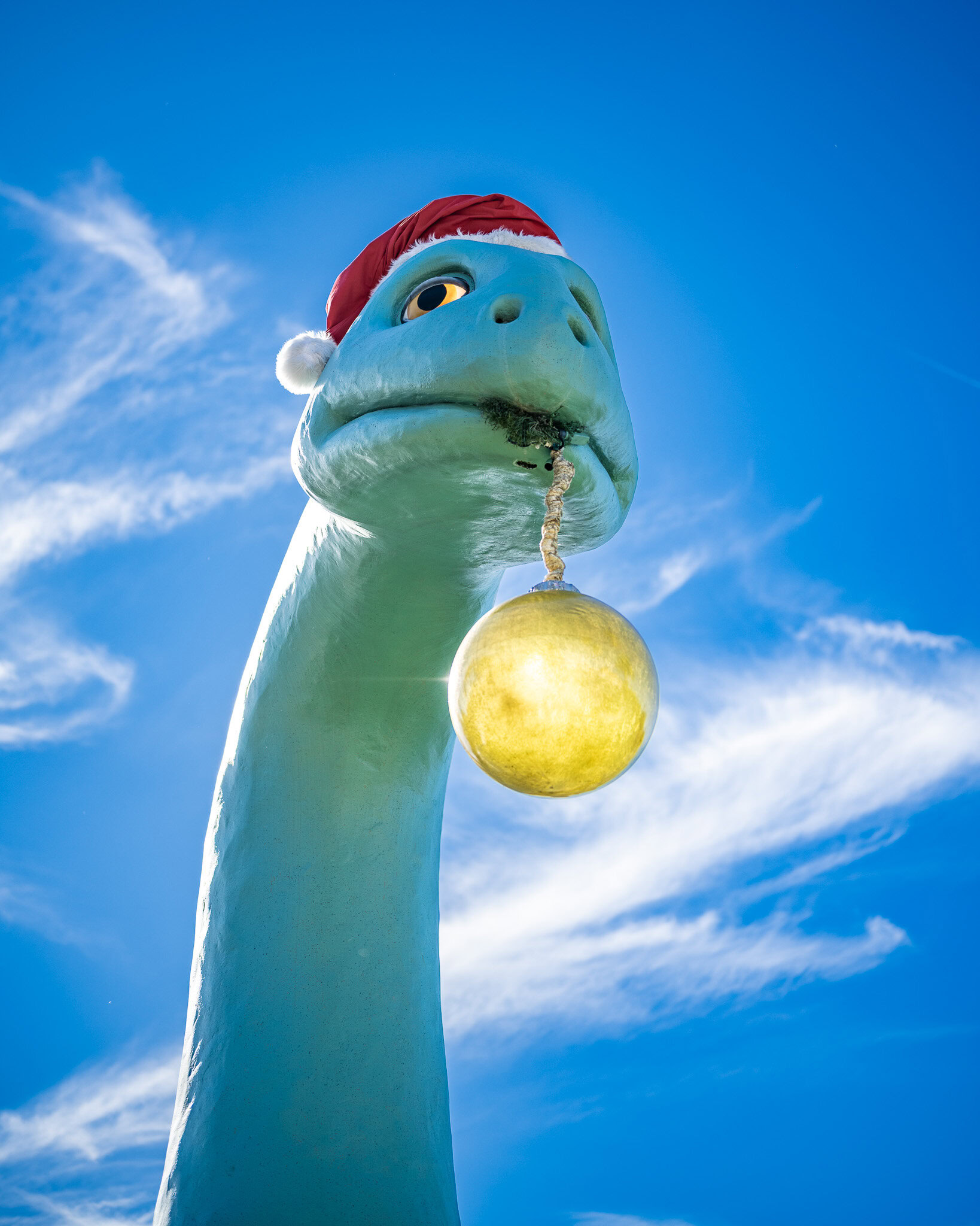 Holiday Decor Dinosaur at Disney’s Hollywood Studios Dinosaur statue at Disney’s Hollywood Studios wearing a Santa hat and holding a gold ornament against a bright blue sky.
