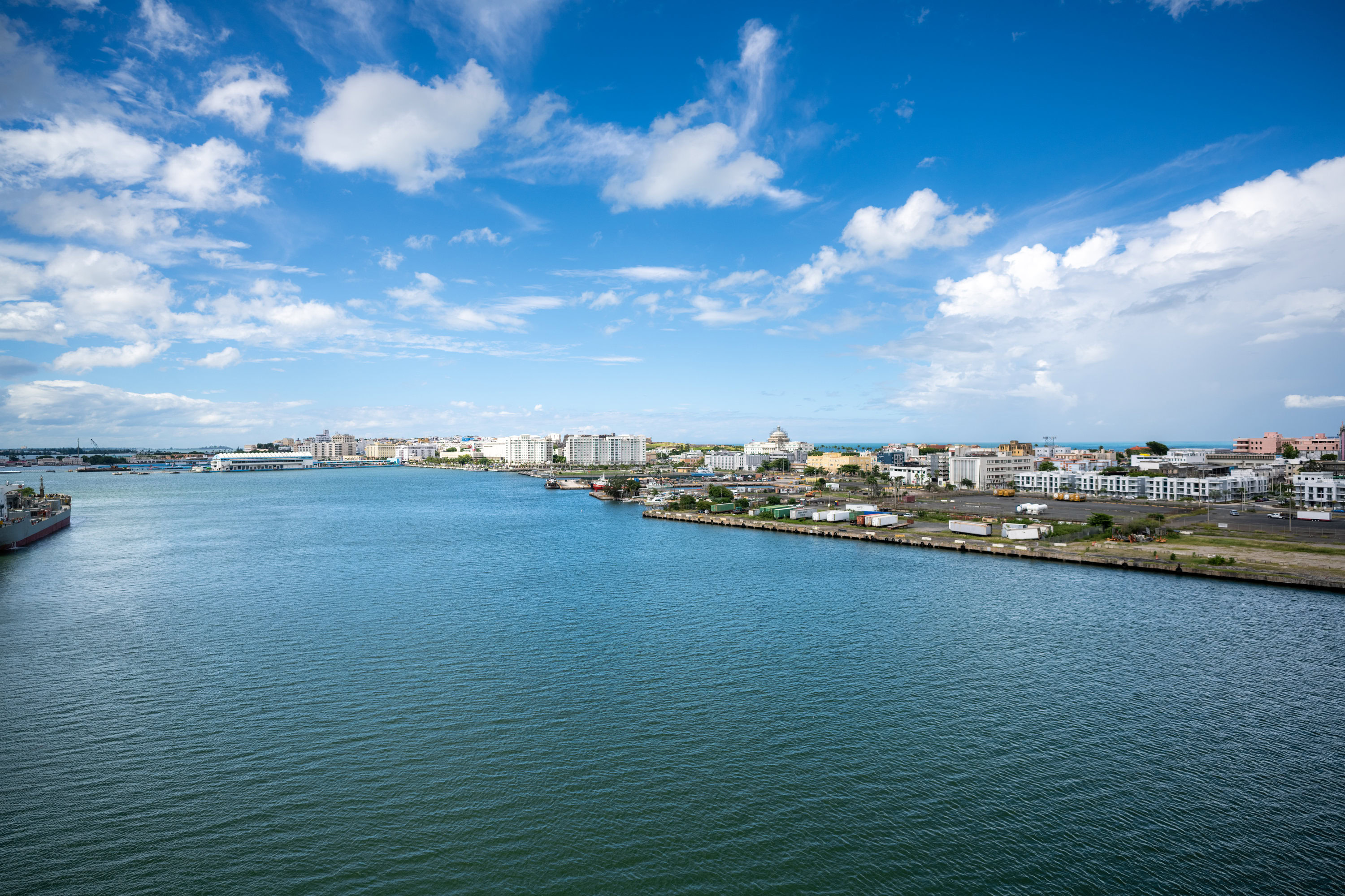 Aerial-style view of Old San Juan, Puerto Rico, taken from the top deck of the Disney Magic cruise ship docked at the Pan American Pier, showing the waterfront, city buildings, and San Juan Bay under a bright blue sky.