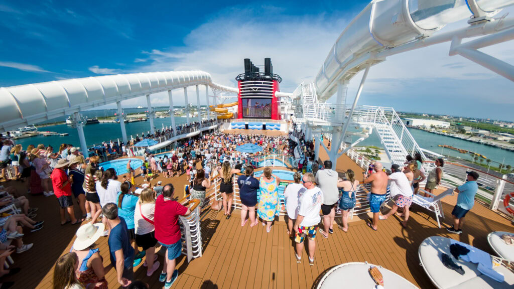 Crowds gathered on the Disney Wish upper deck during the Sail Away Party with AquaMouse slides and the ship&rsquo;s funnel in the background.