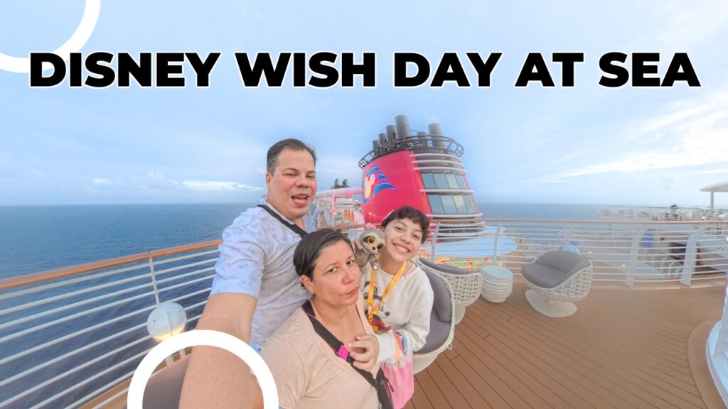 A smiling family taking a selfie on the deck of the Disney Wish cruise ship during a calm sea day, with the ship&rsquo;s signature red funnel and ocean horizon in the background.