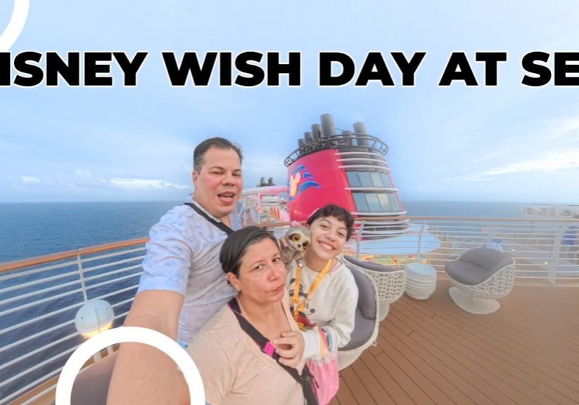 A smiling family taking a selfie on the deck of the Disney Wish cruise ship during a calm sea day, with the ship’s signature red funnel and ocean horizon in the background.