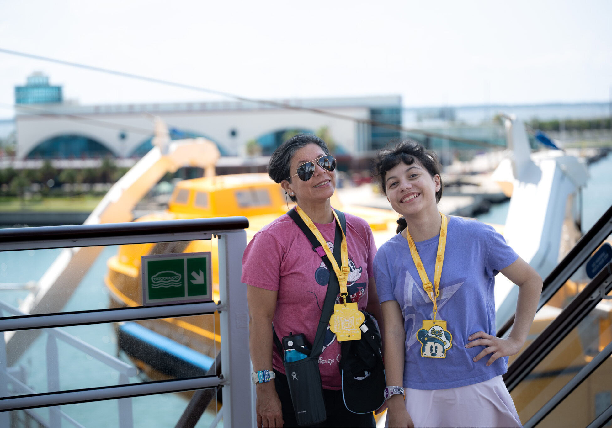 Lucy and Daniela from Laugh and Gear smiling aboard the Disney Wish cruise ship at Port Canaveral, wearing Disney lanyards, with the cruise terminal and a yellow tender boat in the background.